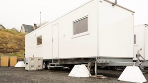 A white mobile home with a small window and a door, parked on a gravel surface with a wooden fence and houses in the background. - Mobile Home Park (Strendur)
