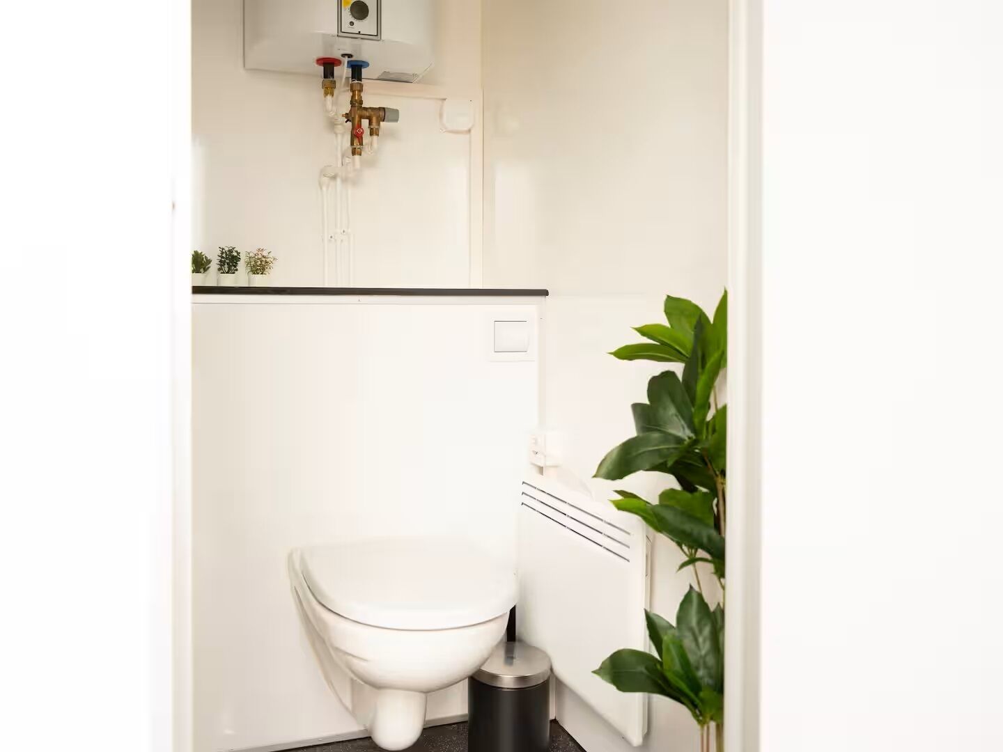 Un baño pequeño y blanco con inodoro, radiador y calentador de agua instalado en la pared.