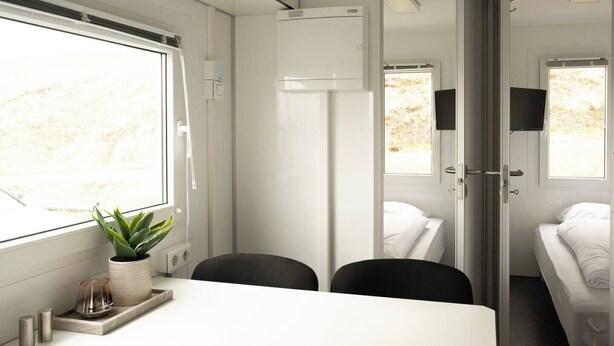 A modern train compartment with a white interior, a small table with a plant, two black chairs, a bed, and a TV mounted on the wall. - Mobile Home Park (Strendur)