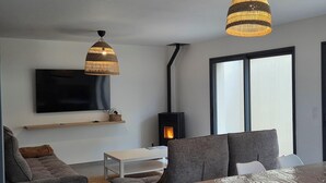 A modern living room with a flat-screen TV, two grey sofas, a wooden dining table, and a black wood-burning stove. - COZY APARTMENT IN FORMIGUERES, SLEEPS 6–8 (Formiguères)