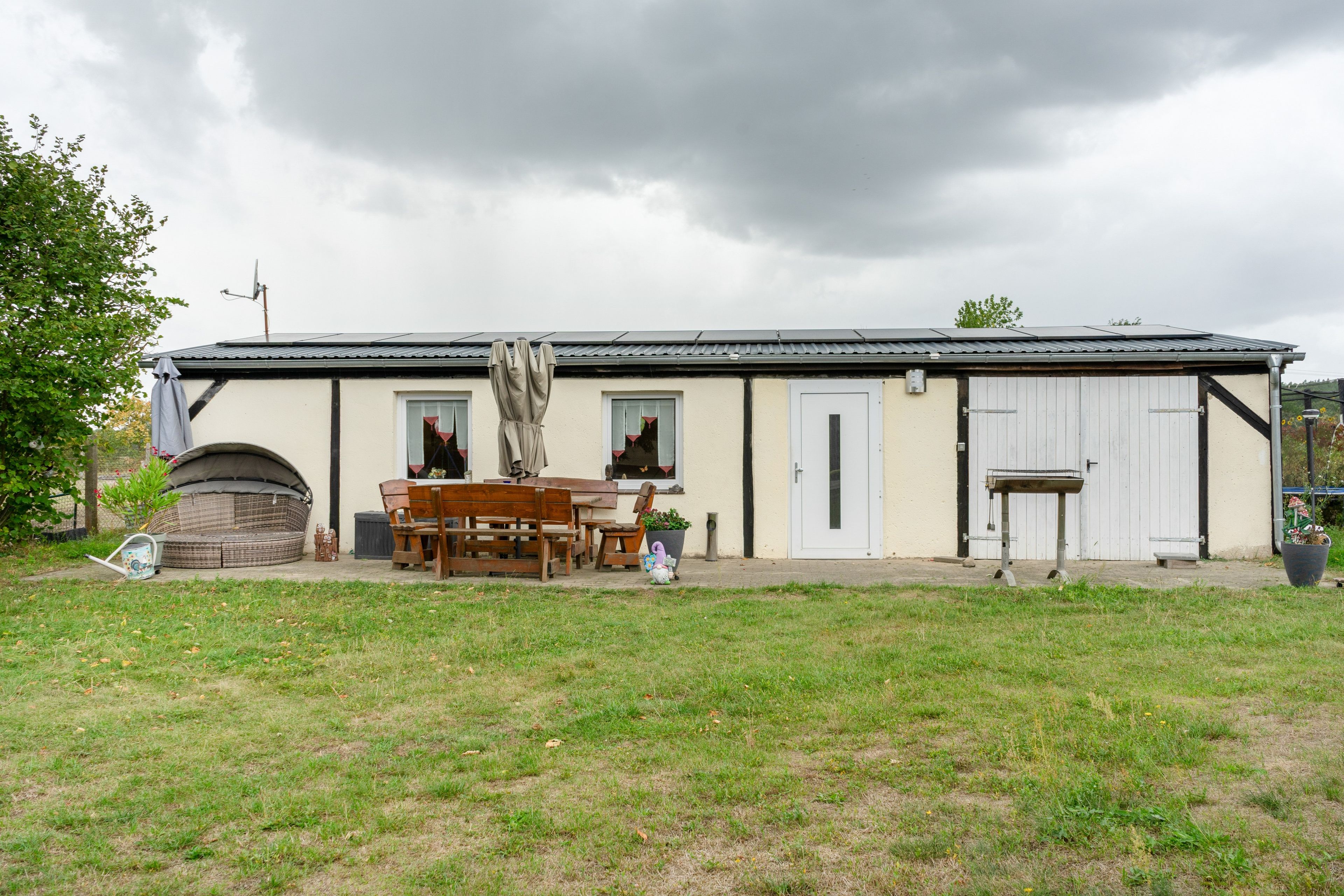 Ein eingeschossiges Gebäude mit einer Terrassenfläche, die einen Holztisch, Stühle, einen Grill und einen Blumentopf zeigt.