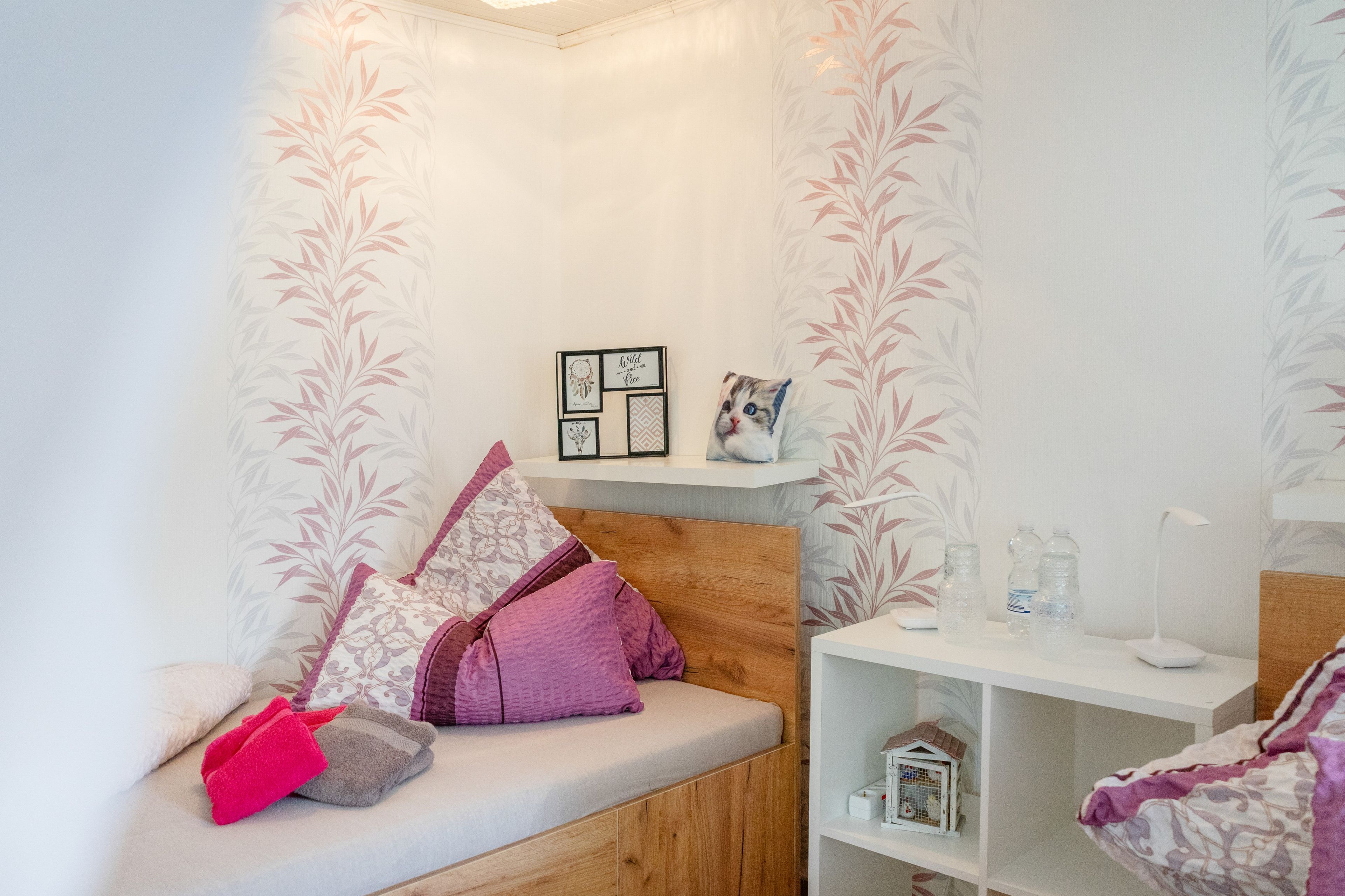 A cozy room with a wooden headboard, a bench with cushions, a shelf with framed pictures, and a small white cabinet with bottles.