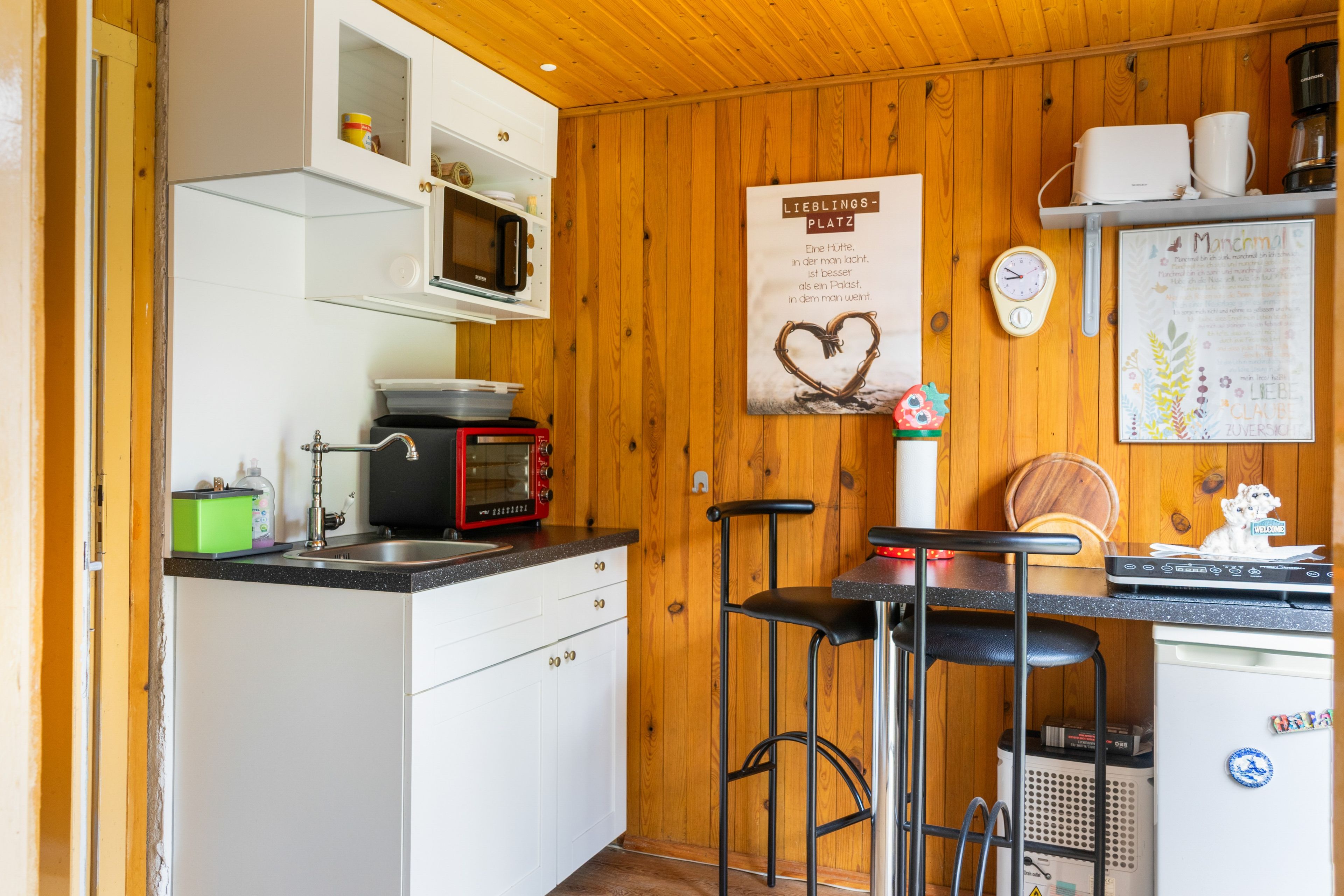 Cozinha compacta com micro-ondas, torradeira e pia. Uma parede de madeira e um cartaz são visíveis.
