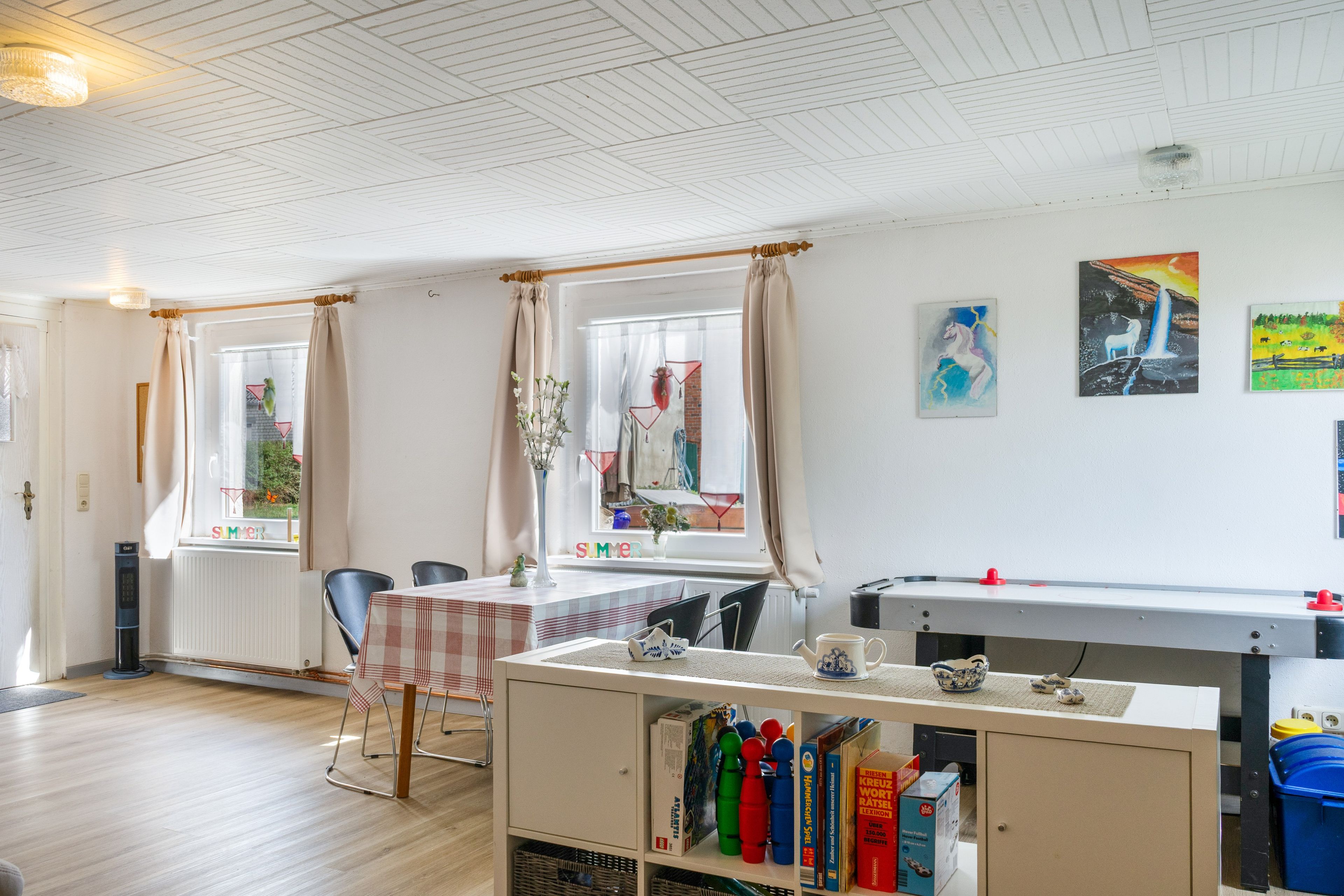 Ein Zimmer mit Holzboden, einem Tisch mit Stühlen, einem Heizkörper und einem Fenster mit Vorhängen.