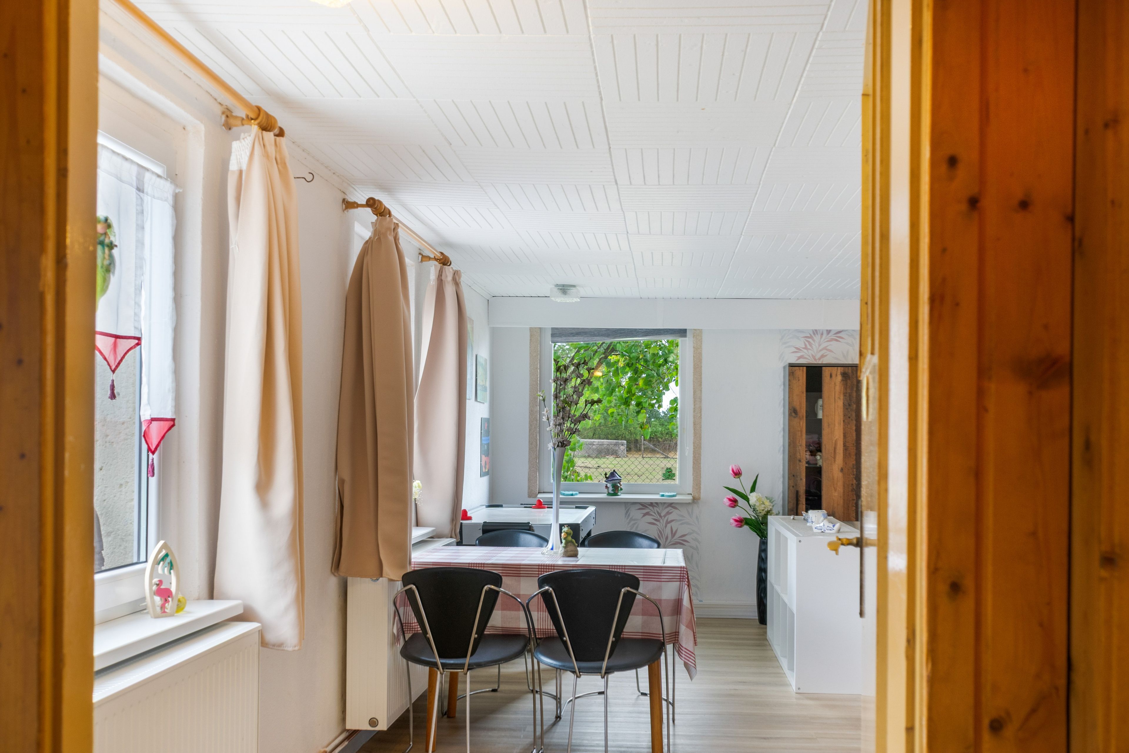 A room with a dining table, chairs, a window with curtains, and a view of greenery outside.