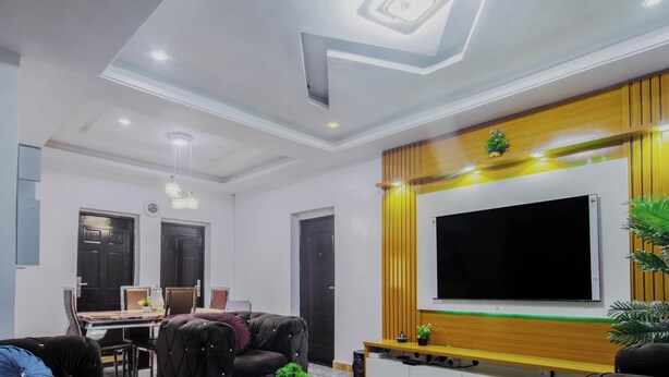 A modern living room with a large flat-screen TV mounted on a wooden wall, a dining area with a table and chairs, and a ceiling with recessed lighting and a decorative design. - Adunola Serviced  Apartment  (Lagos)