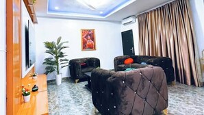 A modern living room with a marble floor, a large brown sofa, a wooden cabinet, a TV, and a painting on the wall. - Adunola Serviced  Apartment  (Lagos)