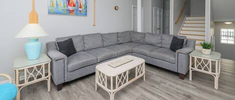 Una sala de estar moderna con un sofá seccional gris, una pequeña mesa de centro blanca y una lámpara azul sobre una mesita auxiliar a juego.