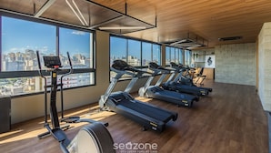 A gym with treadmills, a stationary bike, and a rowing machine, with large windows offering city views. - Condominium w/ Complete Leisure Facilities and Unbeatable Location in POA - Bles (Porto Alegre)