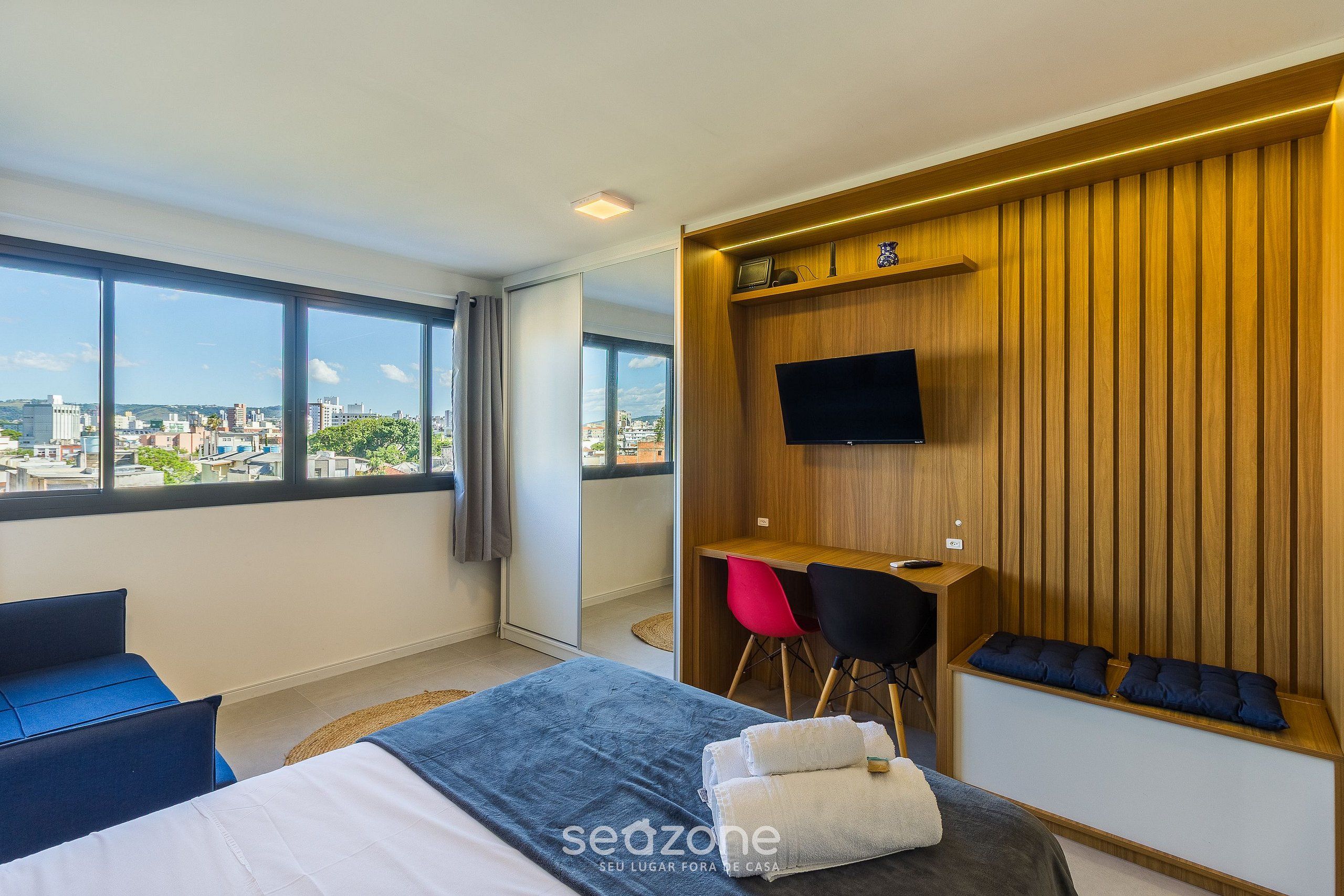 Une chambre d’hôtel moderne dotée d’un grand lit, d’un bureau avec deux chaises, d’un téléviseur fixé au mur et offrant une vue sur la ville grâce à de grandes fenêtres.