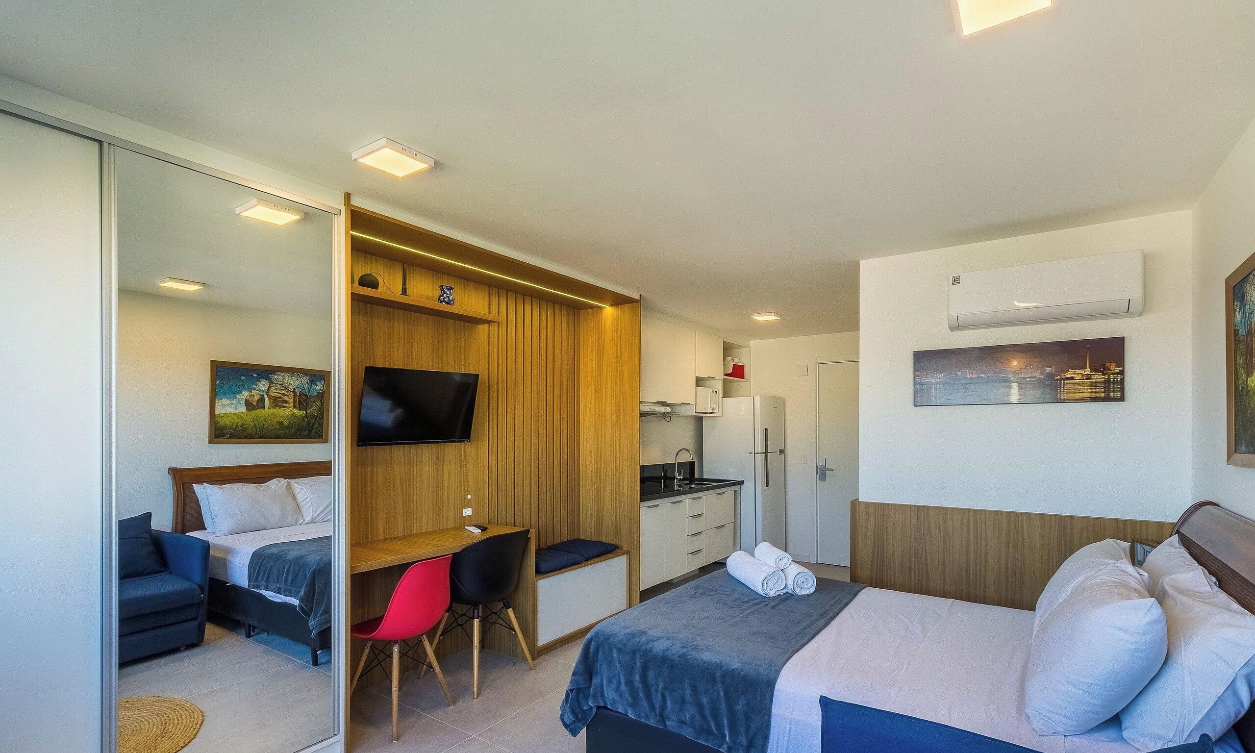 Quarto de hotel com cama, escrivaninha, cadeira, televisão, cozinha e obras de arte nas paredes.
