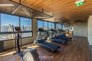 A gym with treadmills, a stationary bike, and a rowing machine, with large windows offering city views. - Condominium w/ Complete Leisure Facilities and Unbeatable Location in POA - Bles (Porto Alegre)