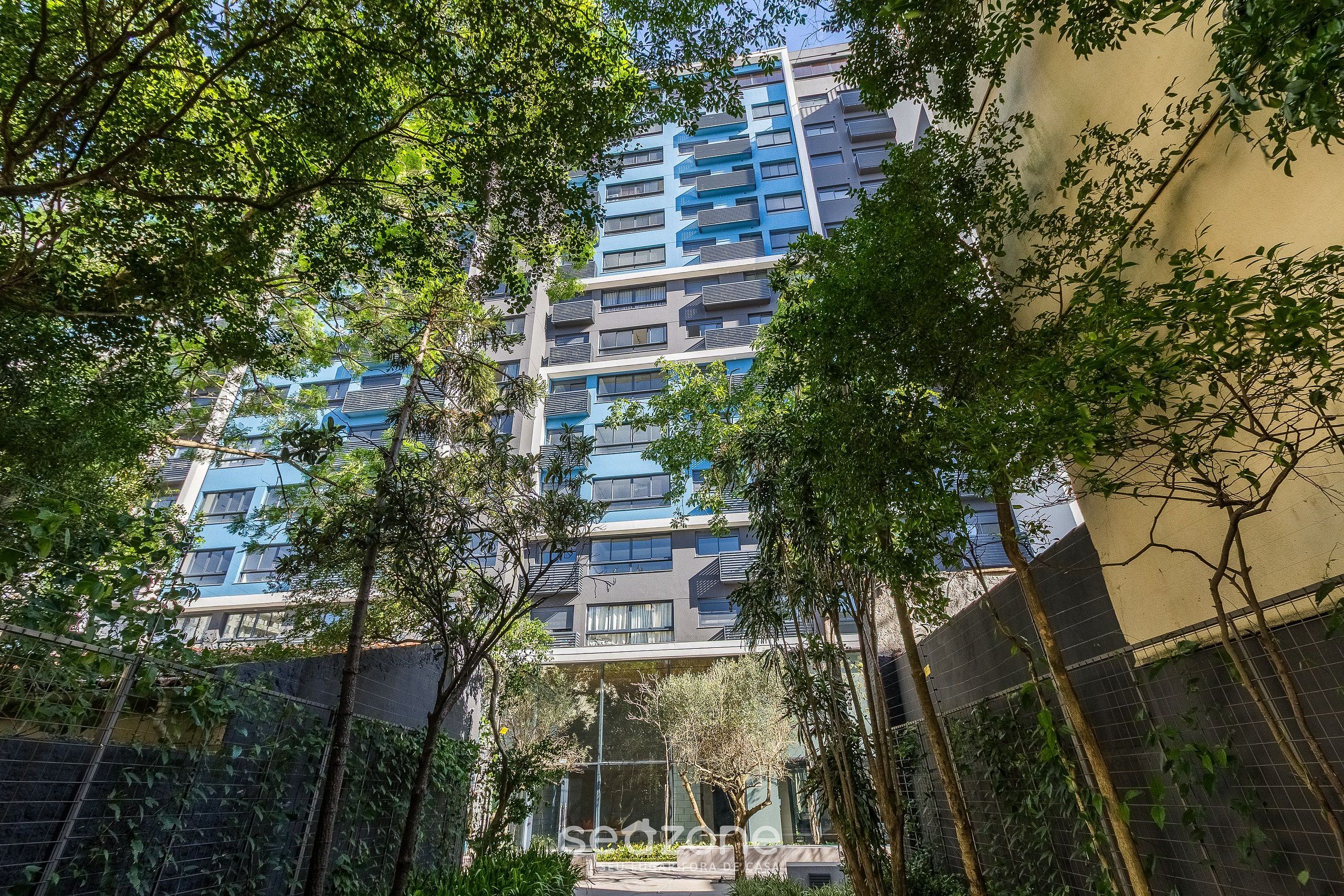 Edifício residencial de vários andares, com varandas, cercado por árvores e um muro.