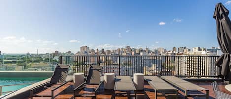 A rooftop terrace with sun loungers, a pool, and a city skyline view.