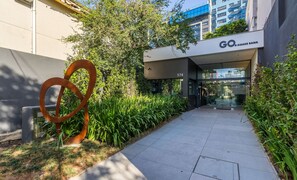 Una entrada moderna a un edificio con una escultura metálica, un camino y abundante vegetación.