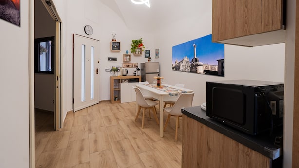 Een gezellige, kleine keuken met een magnetron, een eettafel met stoelen, een klein kastje en een klok aan de muur.