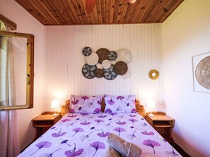Ein Schlafzimmer mit Holzbalkendecke, einem Bett mit violett-blumigem Bettbezug, zwei Nachttischen mit Lampen und Wanddekoration.