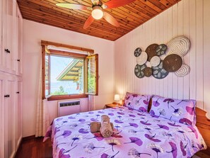 A bedroom with a wooden ceiling, a bed with a purple floral bedspread, a window, and a wall-mounted decorative plate. - Bella Viesta by Interhome (Albisano)