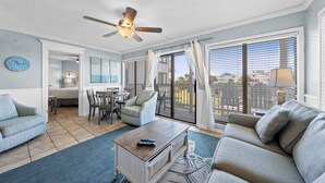 A living room with a sofa, a coffee table, and a ceiling fan. There is a view of the outside through large windows. - GS Plantation West - RCC -1254 (Gulf Shores)