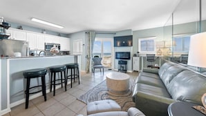 Una cocina moderna con armarios blancos, una isla central con taburetes, un área de comedor con un sofá y una televisión, y un balcón con vistas.