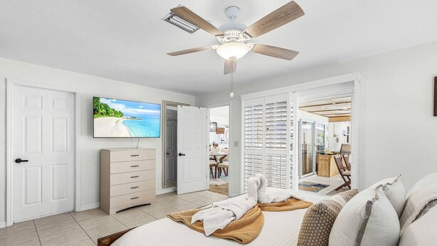 Une chambre à coucher comprenant un lit, un ventilateur de plafond, une télévision, une armoire et une porte coulissante donnant accès à une salle de bain.