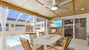 Un coin repas lumineux, donnant sur la piscine, avec des chaises en bois, une table recouverte d’une nappe blanche et un ventilateur de plafond.