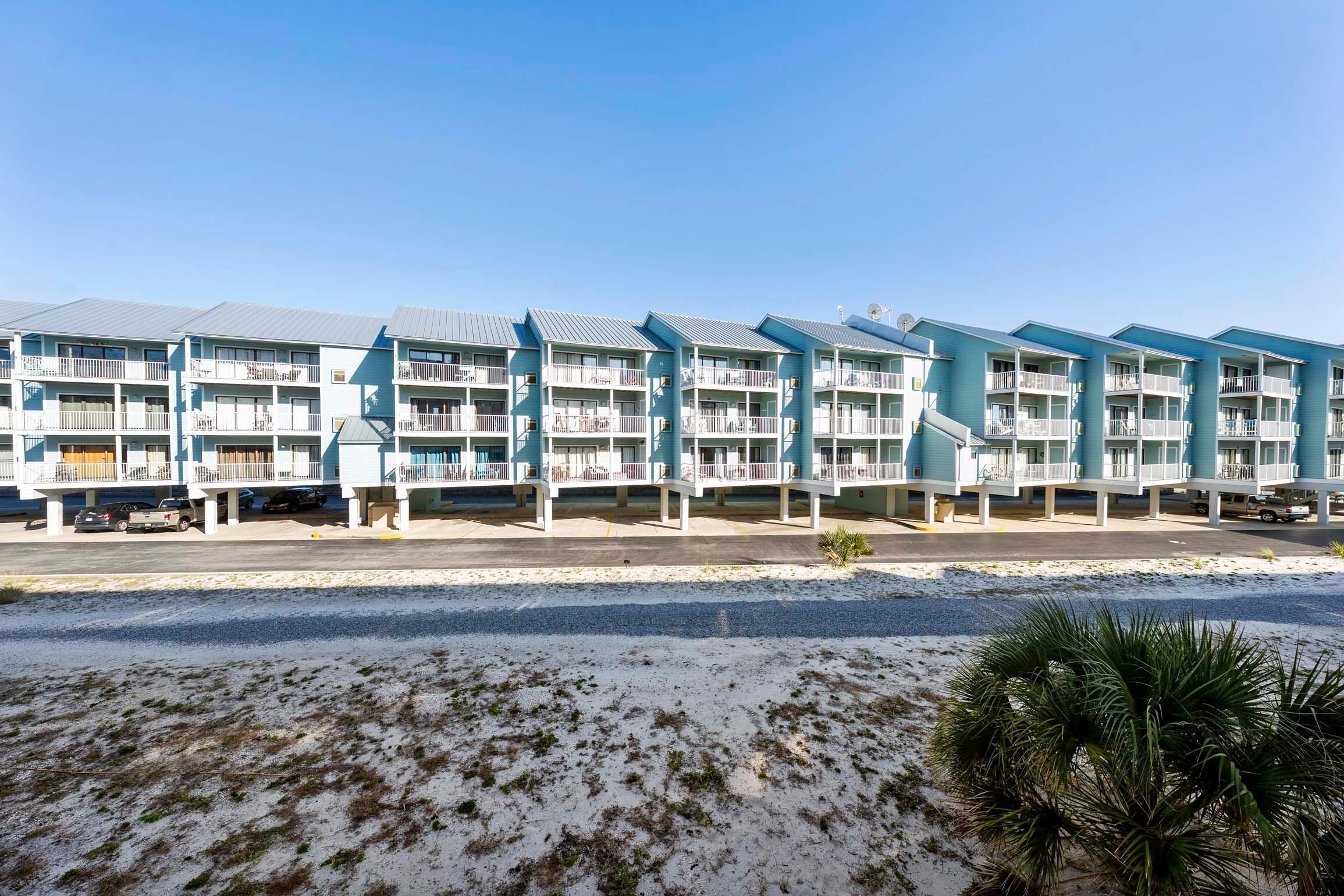 A row of light blue multi-story buildings with balconies, a parking area in front, and a sandy beach area with sparse vegetation.