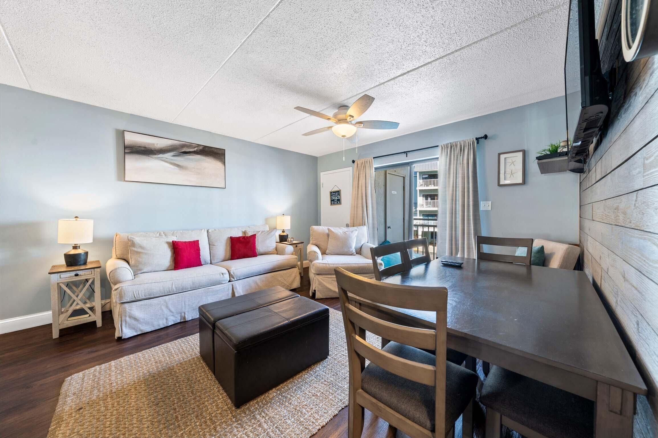 A spacious living area with a ceiling fan, two sofas, a dining table, and a large window with curtains.