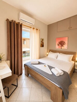 A hotel room with a bed, a desk, a chair, a window with curtains, an air conditioning unit, and a painting on the wall. - Villa Morea With Pool Near Chania City (Mournies)
