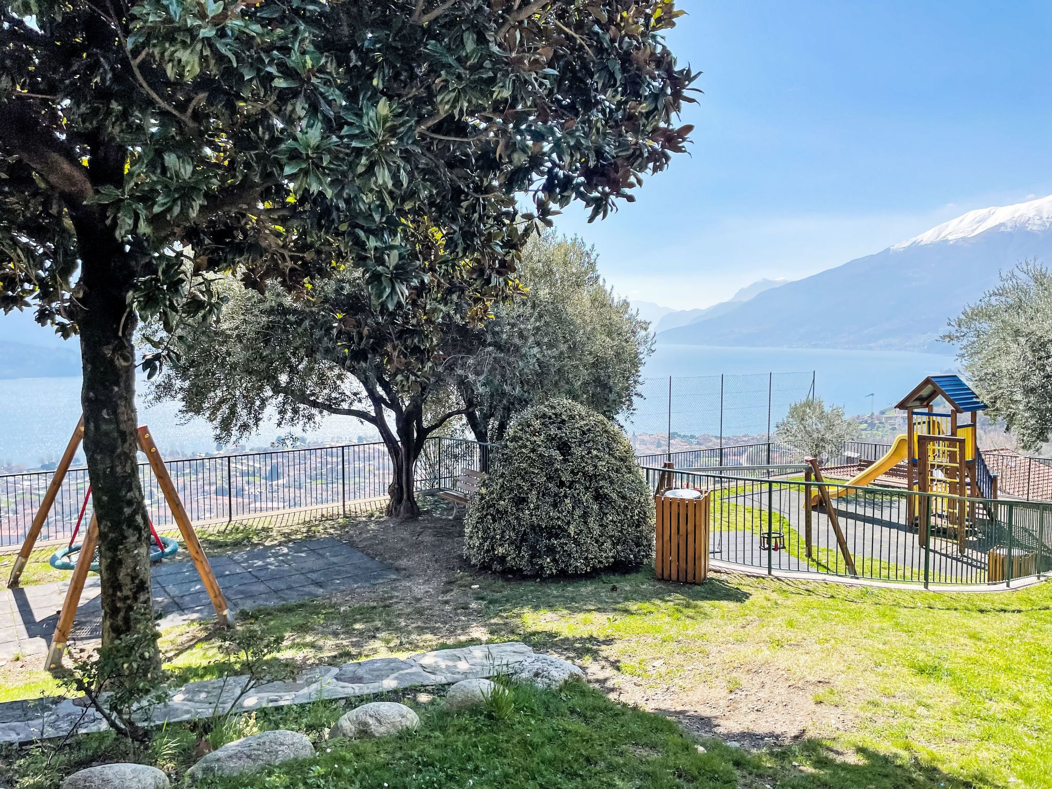 A playground with a slide and climbing structures, surrounded by a fence, with a view of a lake and mountains in the background.