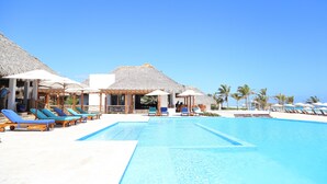 Un espace aménagé au bord de la piscine, avec des chaises longues et des parasols, un ciel d’un bleu limpide et un bâtiment à toit de chaume.