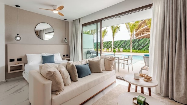 Une chambre d’hôtel moderne dotée d’un grand lit, d’un canapé, d’un coin repas et offrant une vue sur une piscine et des palmiers.