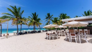 Un complexe hôtelier en bord de mer, doté de chaises longues et de parasols, de palmiers et offrant une vue sur un ciel bleu azur.