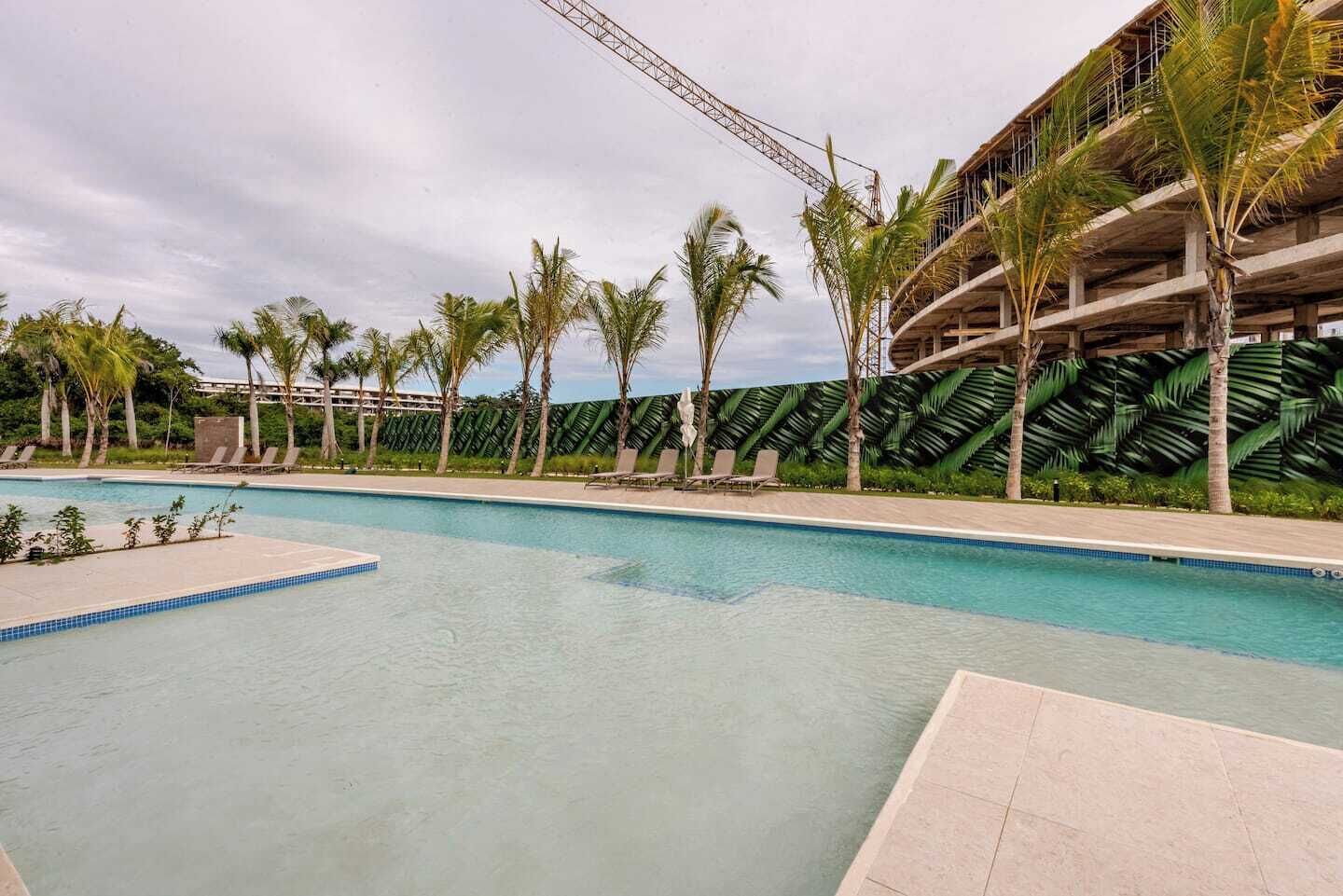 Une piscine entourée de chaises longues et de palmiers, le tout bordé par un bâtiment à la façade ornée de motifs verts.