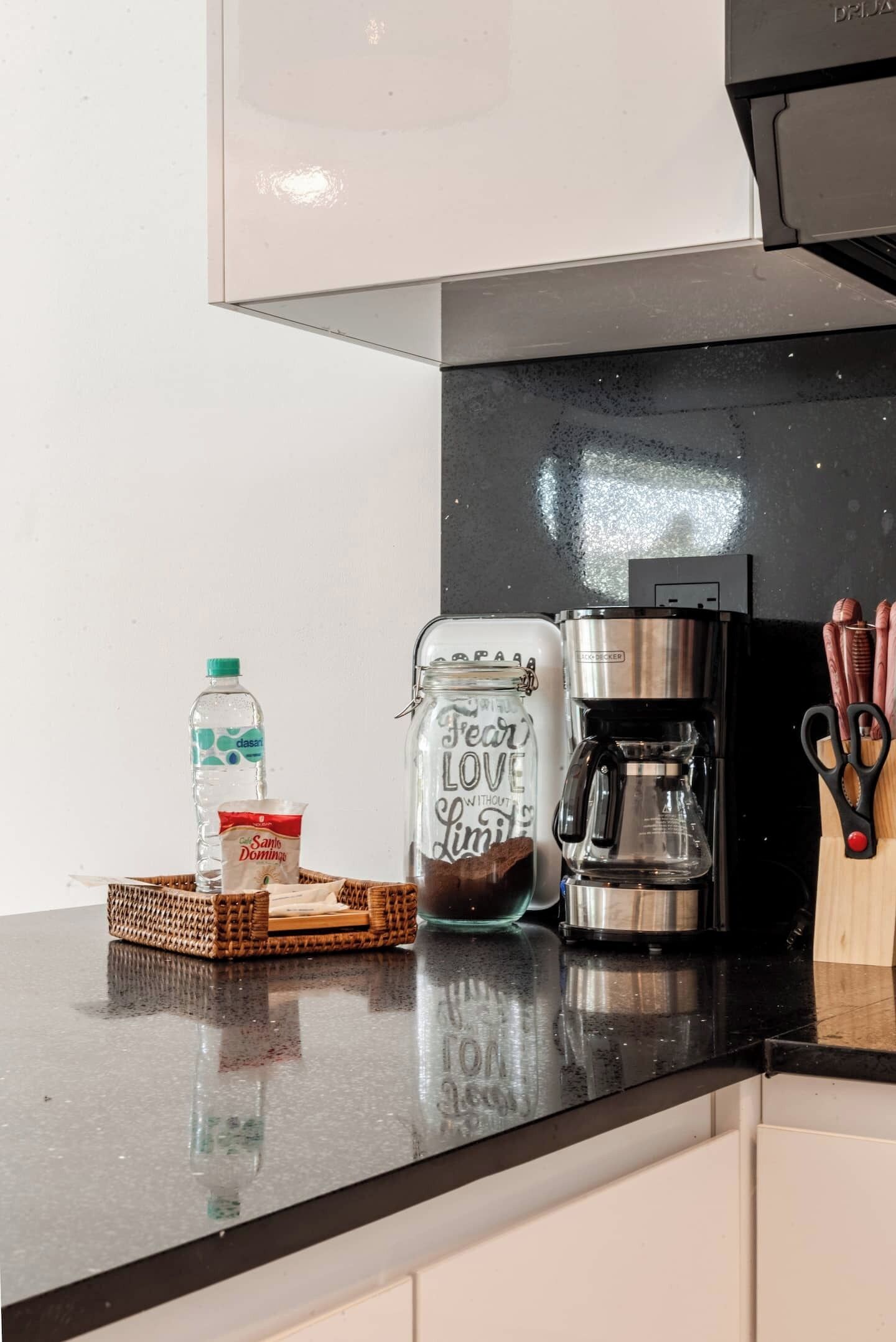 A kitchen counter with a coffee maker, a jar with a message, a water bottle, and a packet of sugar.