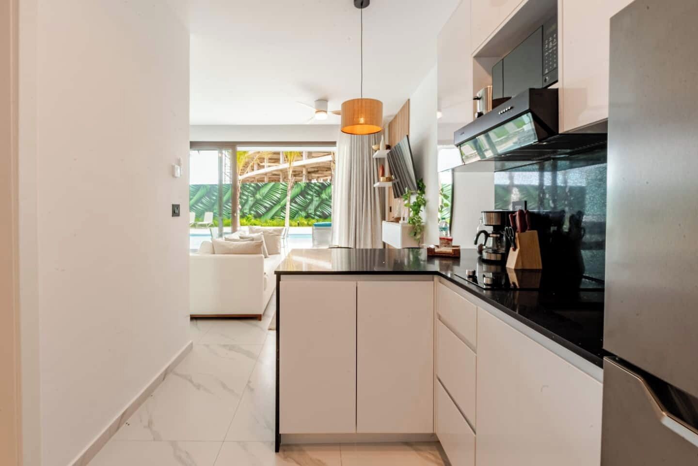 A modern kitchen with a black countertop, white cabinets, and a built-in oven. There is a dining area with a white sofa and a view of a lush garden.