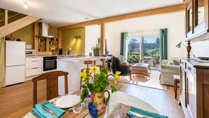 Eine geräumige Küche mit Holzbalken, ein Essbereich mit Tisch für zwei Personen und ein Ausblick ins Freie durch große Fenster.