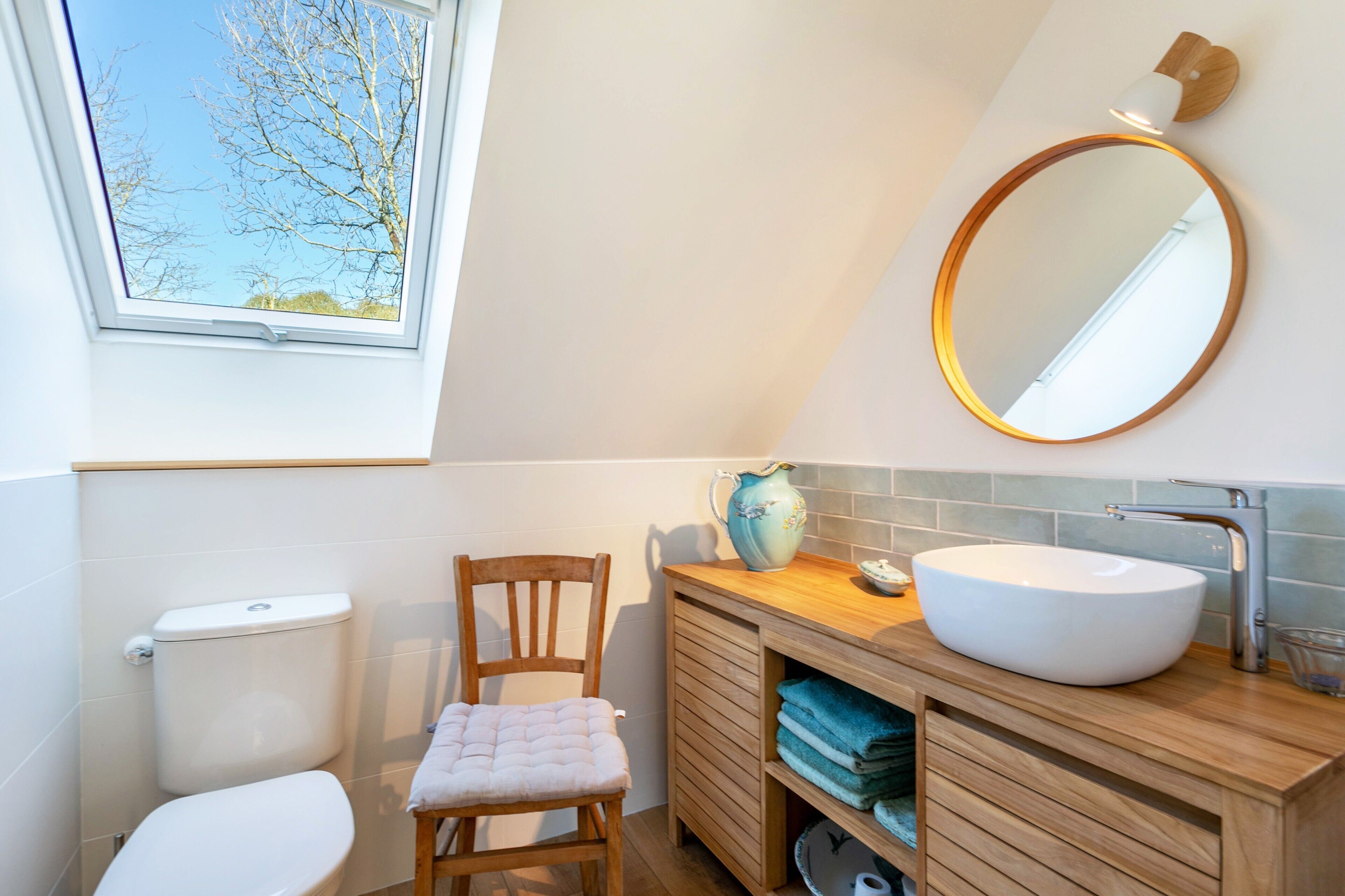 Une salle de bain avec des toilettes, une chaise en bois, un lavabo avec un miroir et une fenêtre.