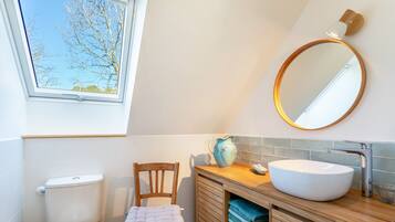 Une salle de bain avec des toilettes, une chaise en bois, un lavabo avec un miroir et une fenêtre.