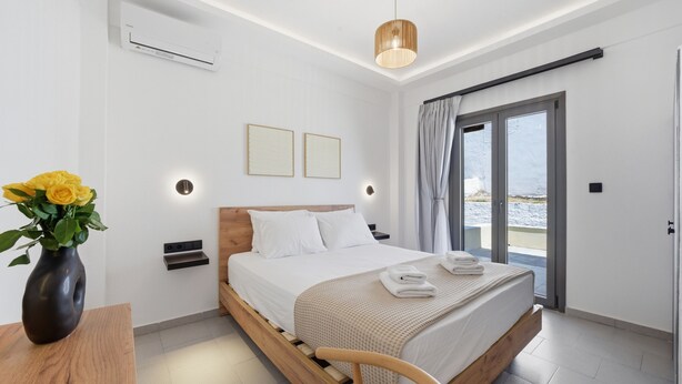 Ein modernes Schlafzimmer mit einem Holzbett, Nachttischen, einer Vase mit gelben Blumen und Meerblick durch eine Glastür.