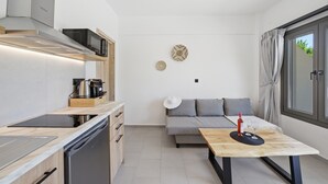 Una cocina compacta con microondas, estufa y lavavajillas, una mesa de comedor con una botella y vasos, un sofá y un ventanal grande.