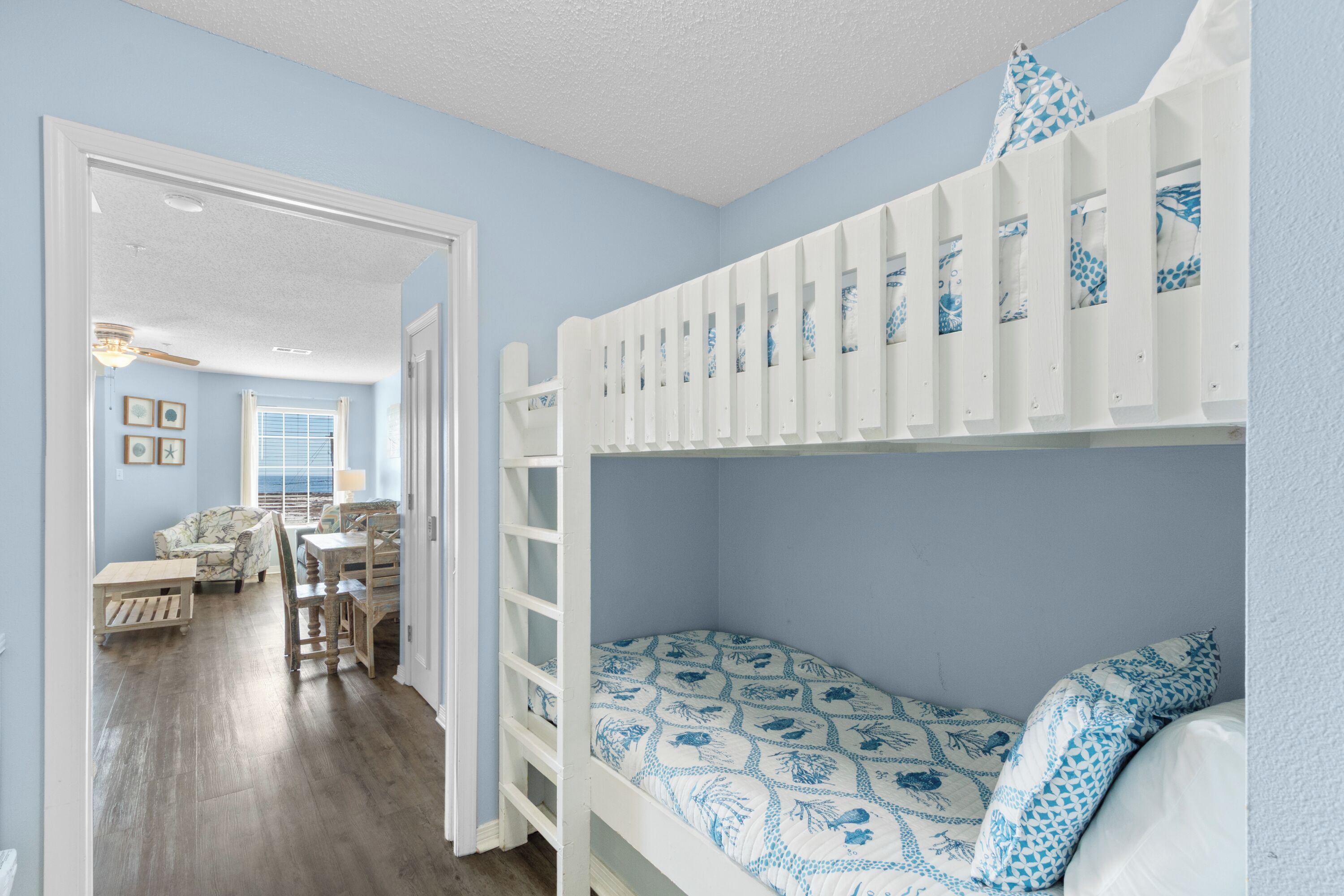 Een slaapkamer met stapelbed, een blauwe muur, houten vloer en uitzicht op een zithoek met een bank en een tafel.