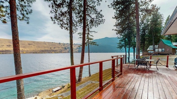 A wooden deck with a red railing overlooking a lake, surrounded by trees and a distant mountain. - Prop House | Wallowa lakefront home w/dock (Joseph)