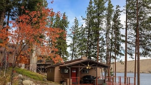 A cabin with a porch, a grill, and a view of a lake surrounded by tall trees. - Prop House | Wallowa lakefront home w/dock (Joseph)