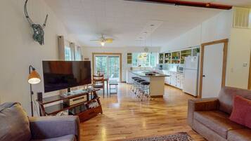A spacious living area with a TV, wooden flooring, a sofa, a coffee table, and a kitchen in the background.
