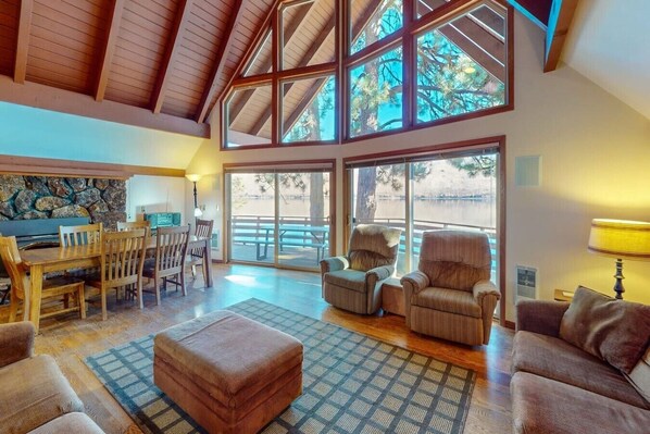 A spacious living area with a high ceiling, large windows, and a stone fireplace.