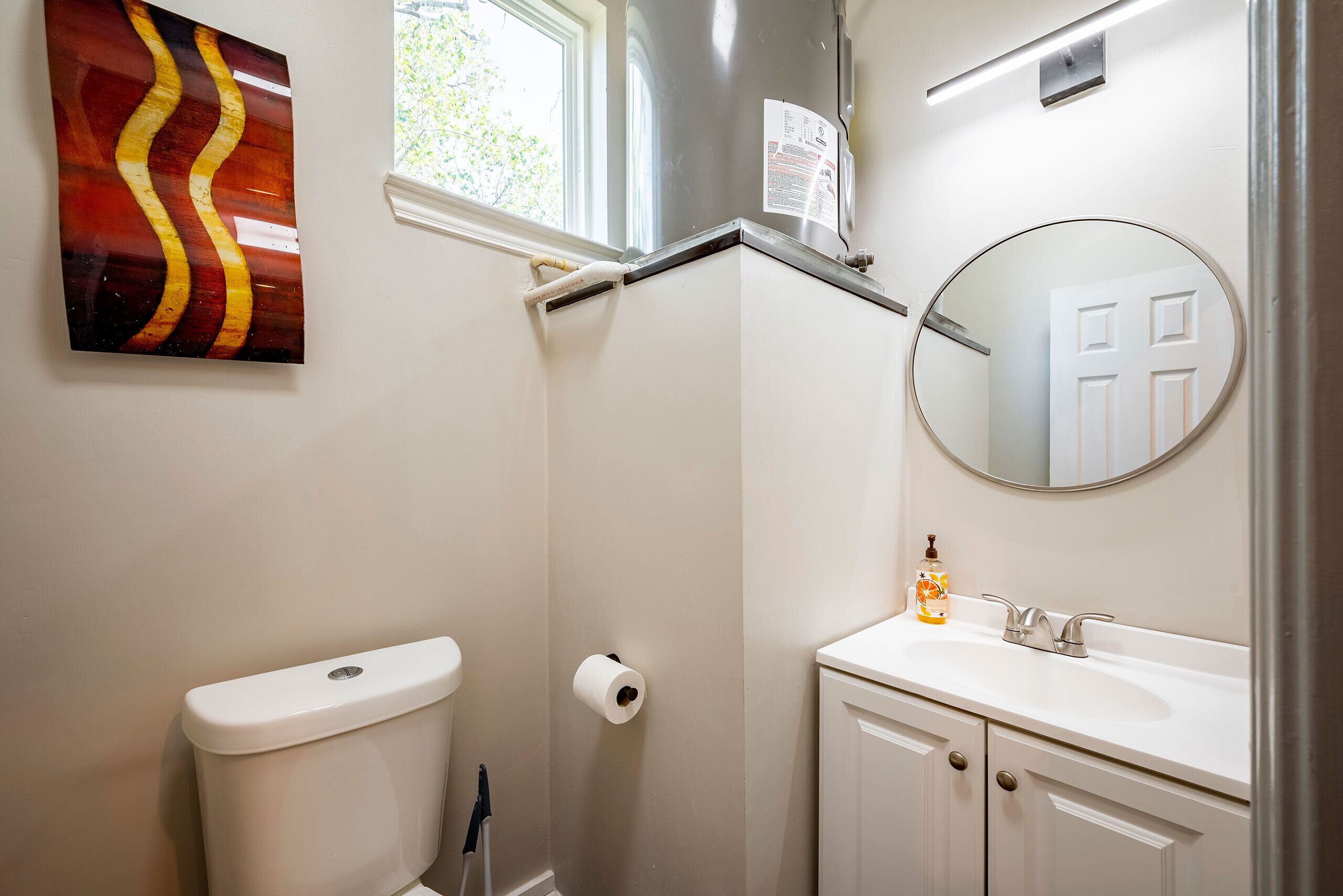 Une salle de bain avec des toilettes, un rouleau de papier toilette, un lavabo avec un miroir et une fenêtre.