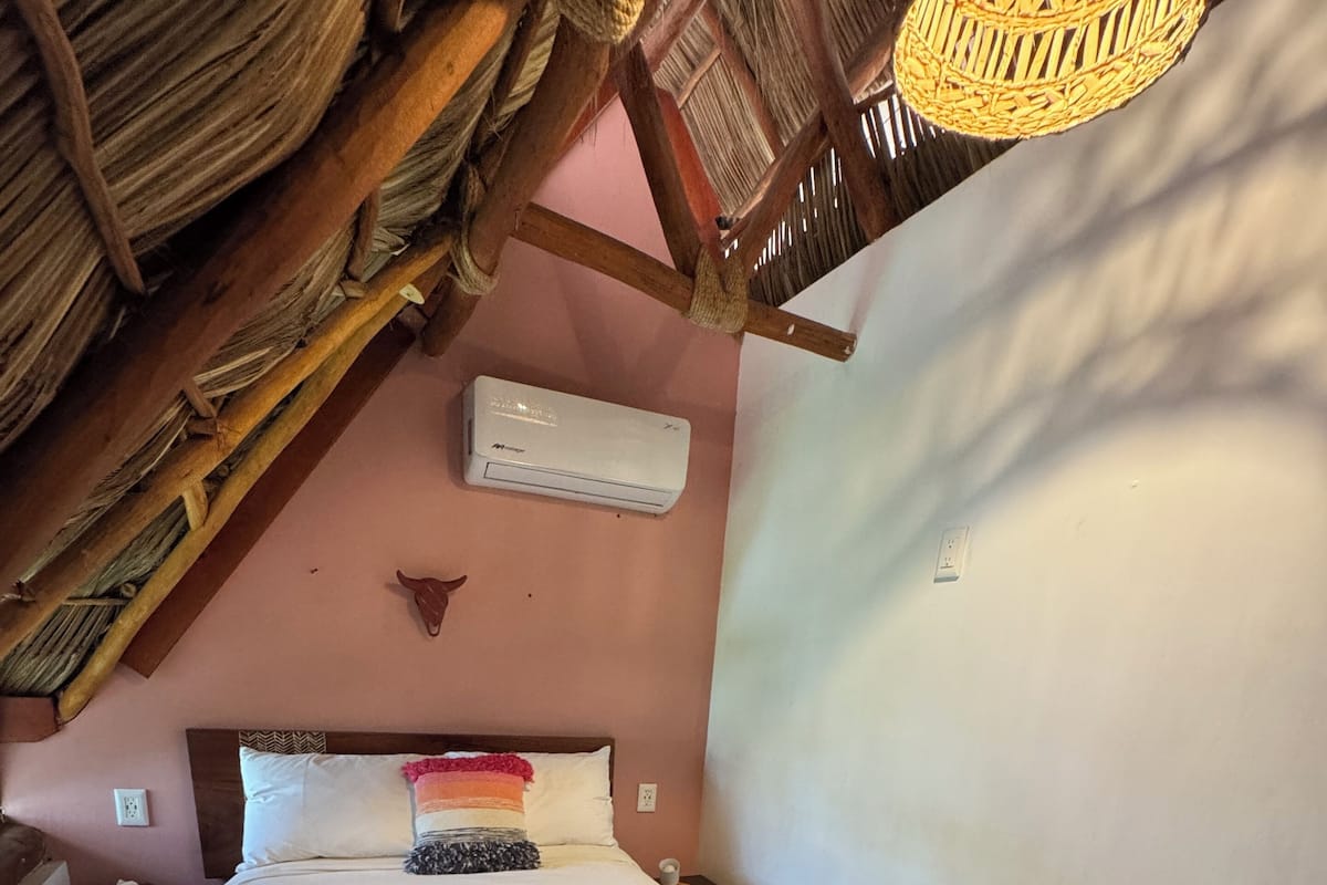 Quarto com teto de madeira, cama com roupa de cama branca e ar condicionado fixado na parede.