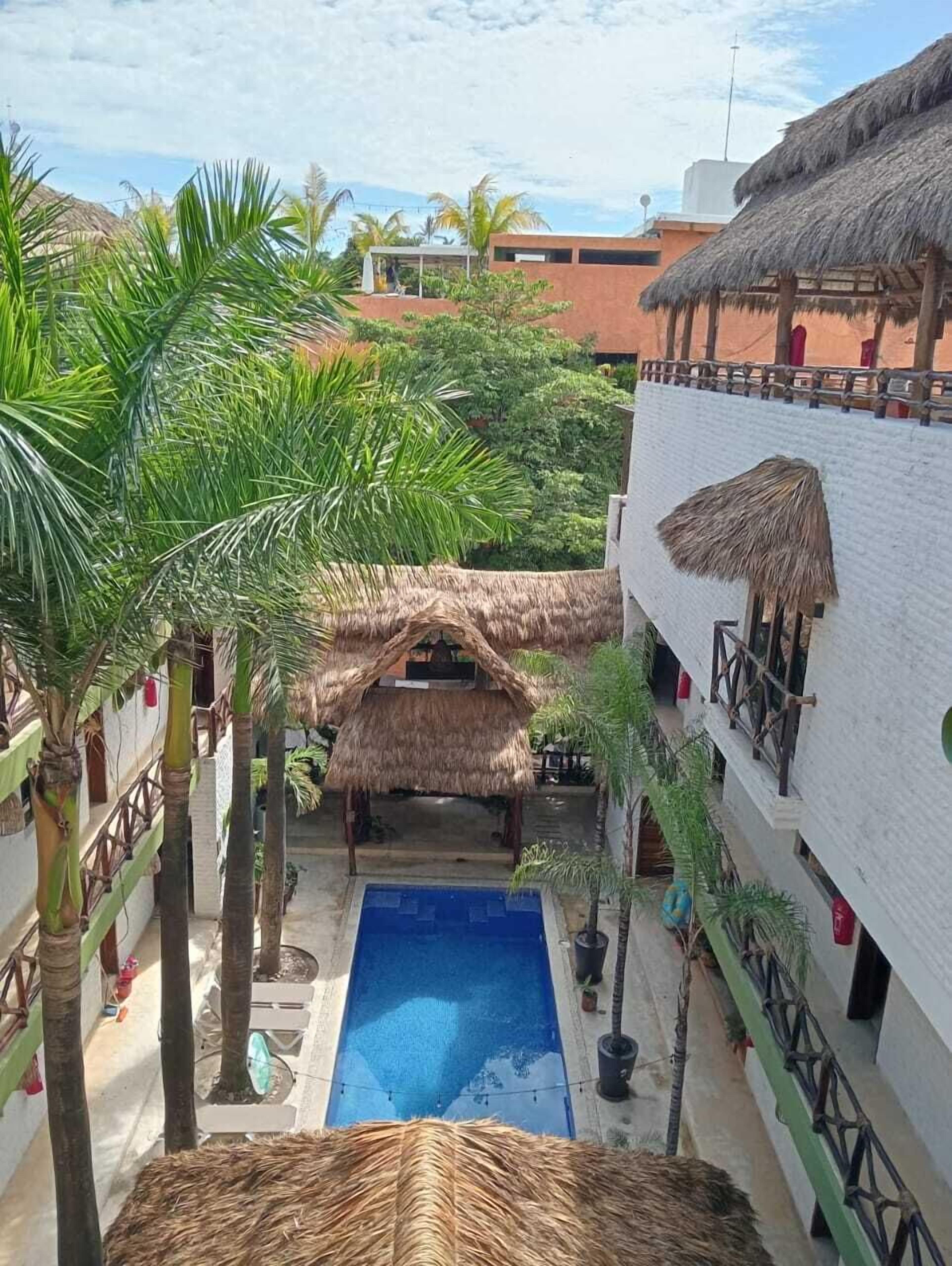 A pool area with a thatched roof structure, surrounded by palm trees and a white building with balconies.
