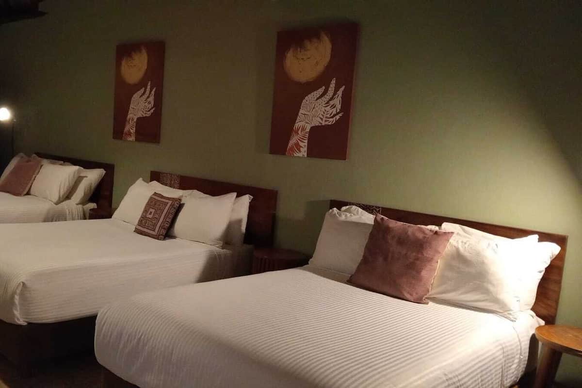 Quarto de hotel com três camas, cada uma com roupa de cama branca e almofadas castanhas. Duas obras de arte penduradas na parede retratam mãos alcançando a lua.